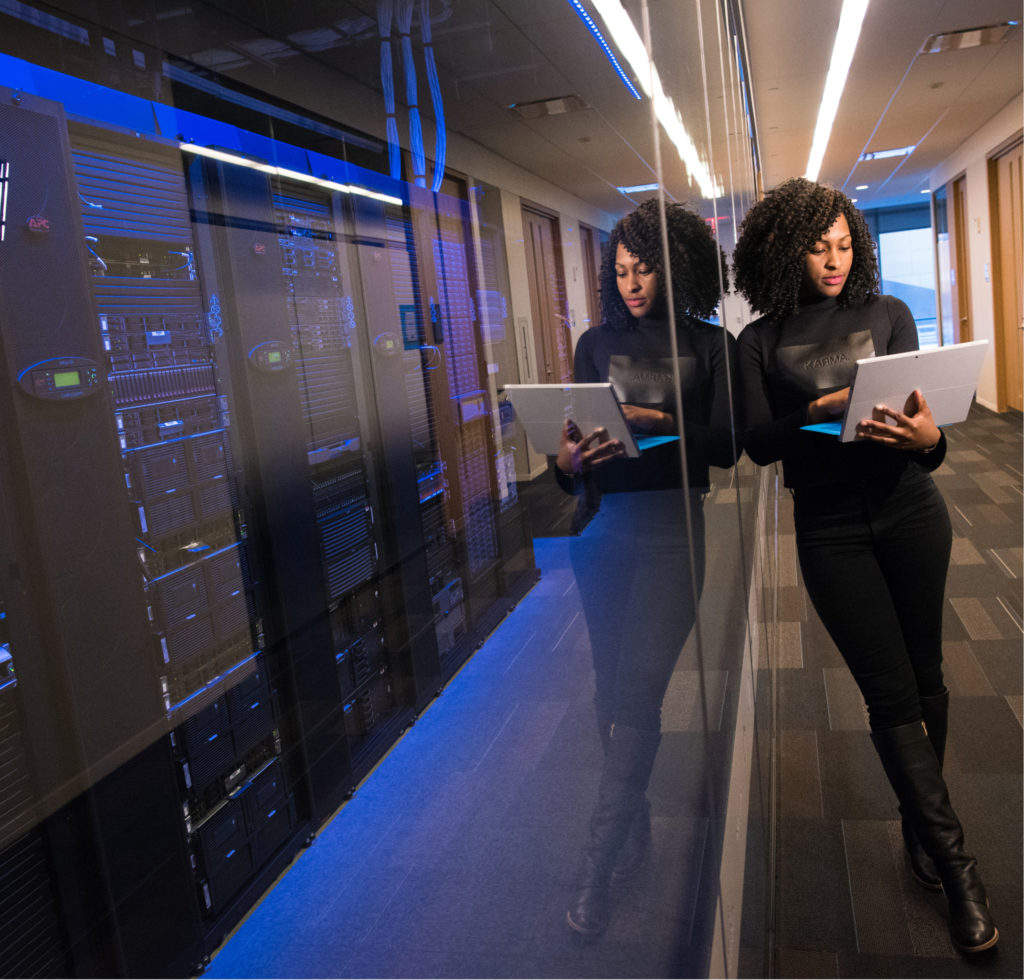 A black woman leans against the glass that is housing black and dark blue illuminated servers. The woman who works at Splunk is holding a laptop and wearing all black clothing. 