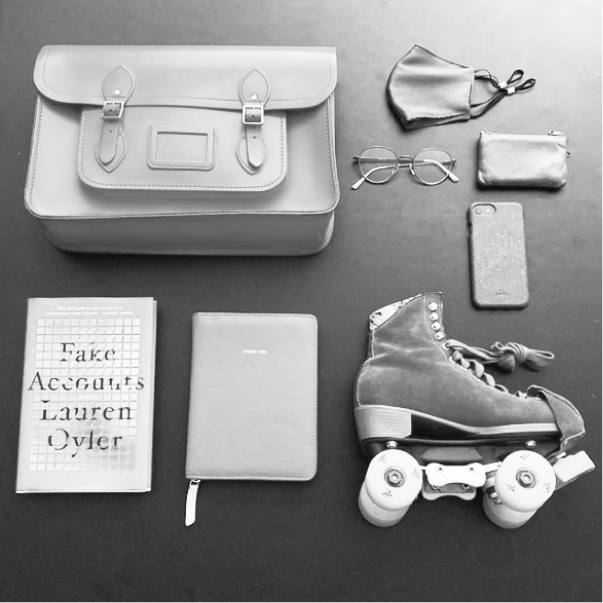 A black and white image of a roller skate, Fake Accounts by Lauren Oyler book, a satchel bag, pair of glasses, mask, iphone, wallet and notes book are laid out against a black backdrop. They are a few of Kathryn L. Hall's favourite things even though she has a career in cloud computing.