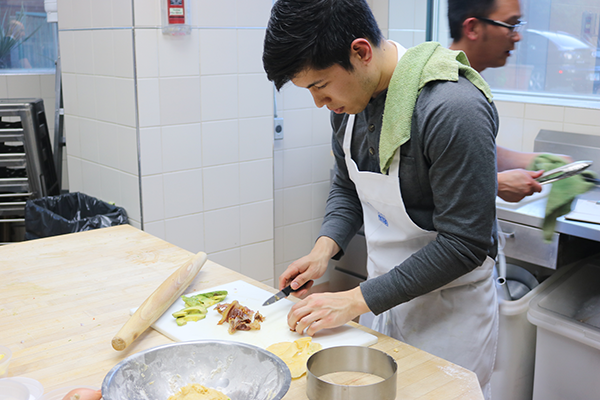 Male Team Member using a knife to chop things.  Hand towel thrown over his left shoulder. A rolling pin and avocado rest near by on the cutting board.