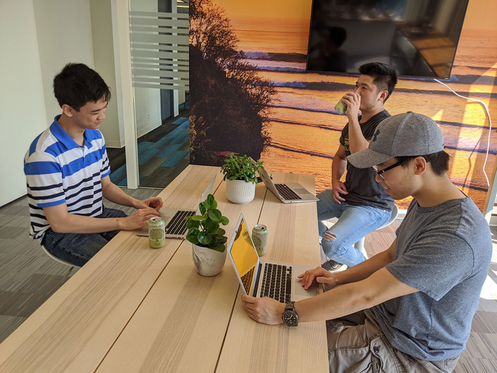 Three interns sit around a light coloured wood table each with a laptop in front of them. There are two plants on the table. From left to right is Bowen, Victor, and Andy. Bowen and Andy smile to themselves as they work on their laptops. Victor has a sip of his cider.
