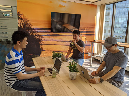 From left to right sits Bowen, Victor, and Andy sitting in the main kitchen around a square table with sunlight streaming in. They are 3 Asian men, Bowen is in a blue and white horizontal striped t-shirt. Victor is taking a sip of his cider as their laptops are open for Friday Retro. Andy and Bowen are laughing.